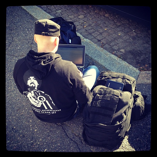 Man with black cap and hoodie sitting in pawement and hacking with a laptop